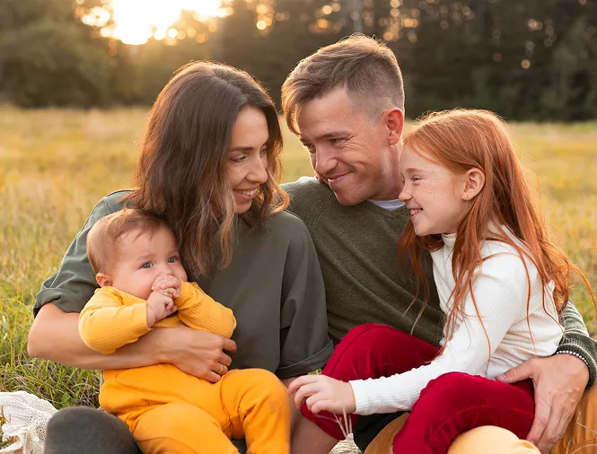 Smiling family of four sitting on grass in a sunlit field, with mother holding a baby and father embracing a young red-haired girl.