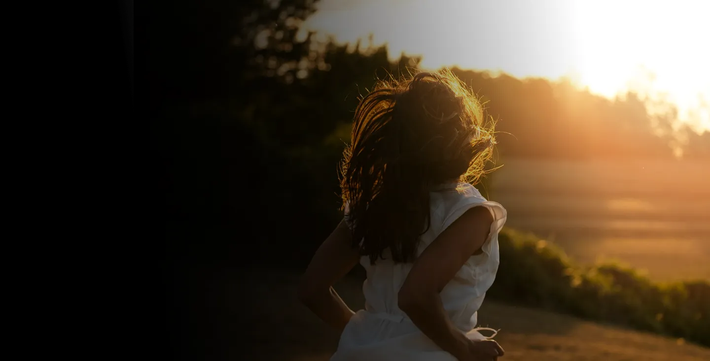 A woman with long hair running outdoors at sunset with sunlight illuminating hair and white dress.