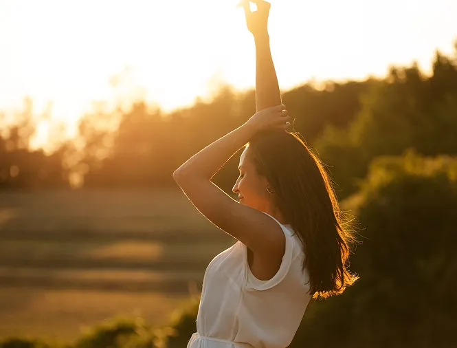 Woman in a white dress raising her arms and enjoying sunlight with blurred outdoor background.