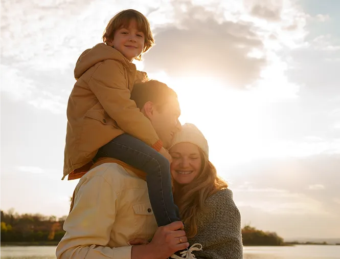 A child wearing a brown jacket sits on an adult's shoulders with another adult hugging them, all outdoors at sunset by the water.