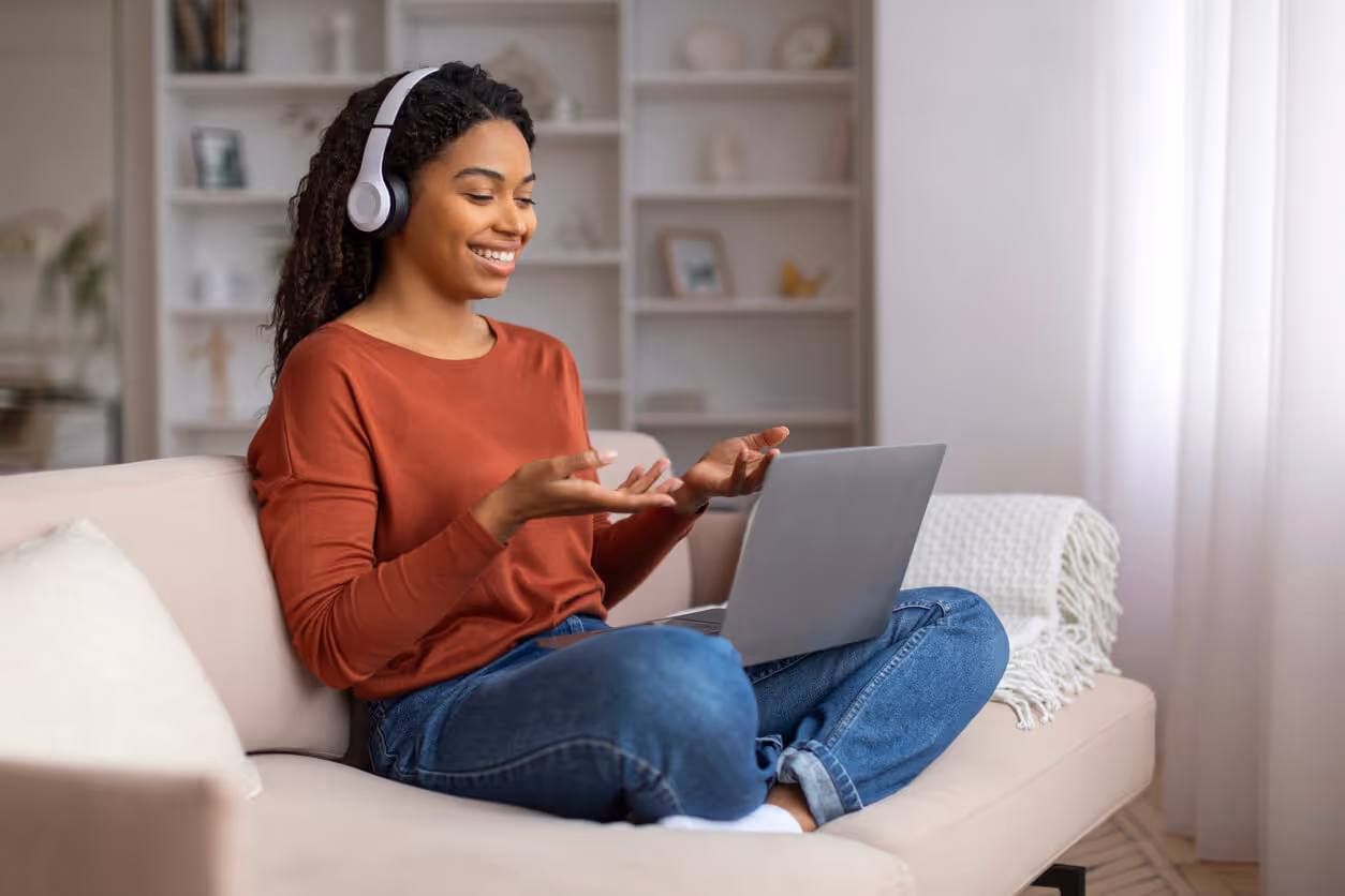 Smiling woman wearing headphones sitting cross-legged on a couch, video chatting on a laptop.