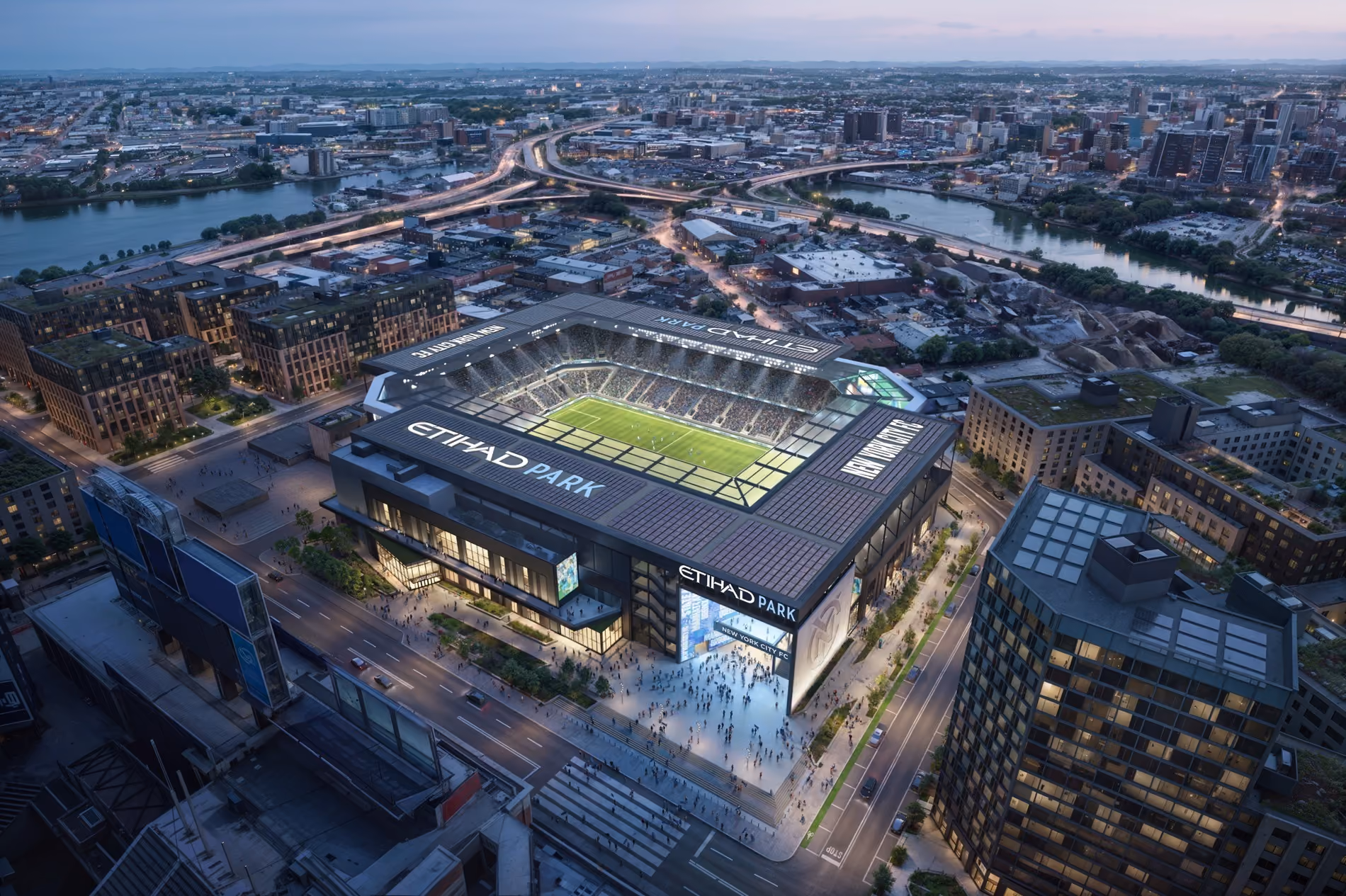 Aerial view of Etihad Park stadium with a soccer match at dusk in an urban cityscape.