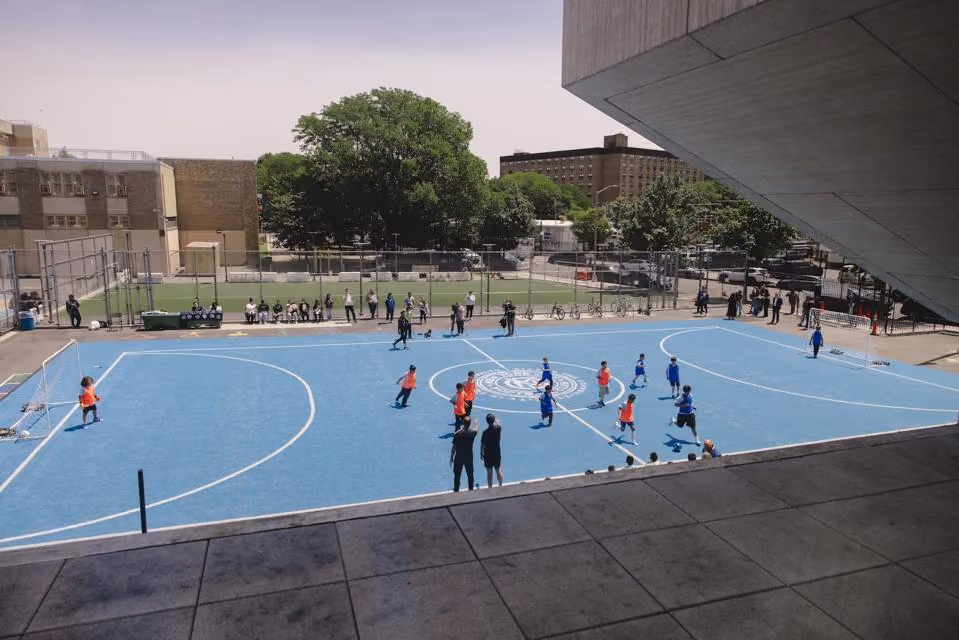 Children playing soccer on a blue outdoor court with coaches and spectators nearby under broad daylight.