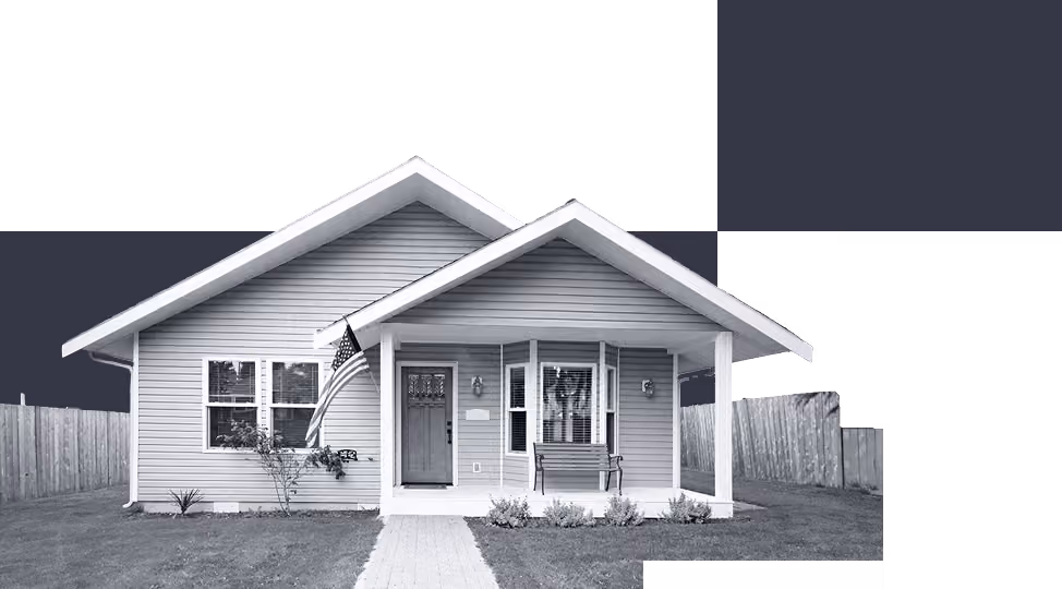 Small single-story house with porch, bench, American flag, and wooden fence on grass lawn.