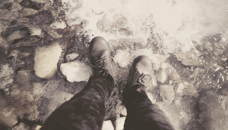 Looking down at boots stepping across rocks in a shallow stream.