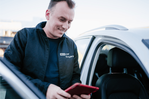 A man wearing a black jacket with the 1800 Car Buyers logo smiles while using a tablet beside a white car with the door open.