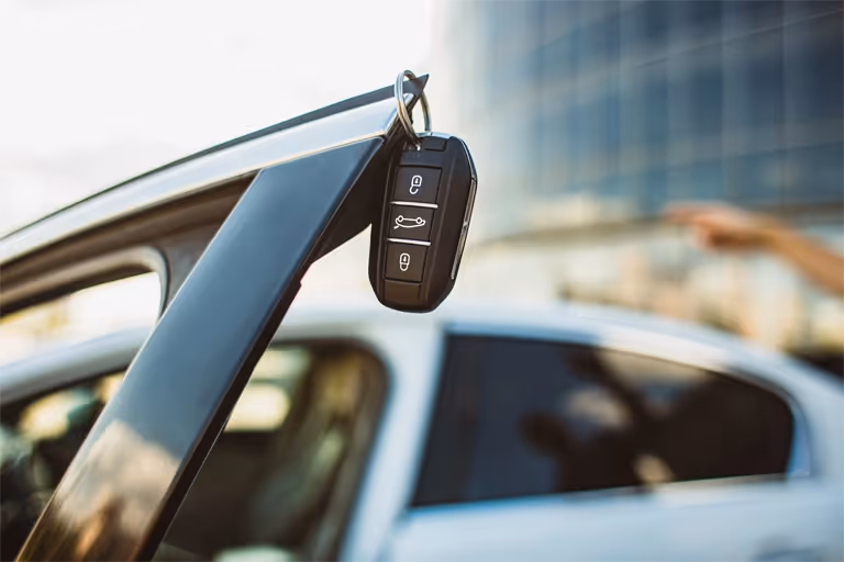 Car key hanging from an open car door with a blurred vehicle in the background.