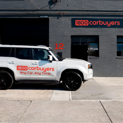 A white SUV featuring orange 1800 Car Buyers branding is parked in front of a grey brick building with matching signage.