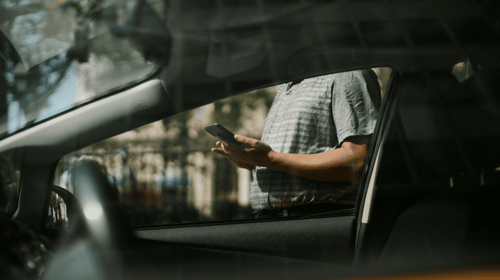 A man holding a mobile phone is viewed through the open window of a car, with reflections visible on the windscreen.