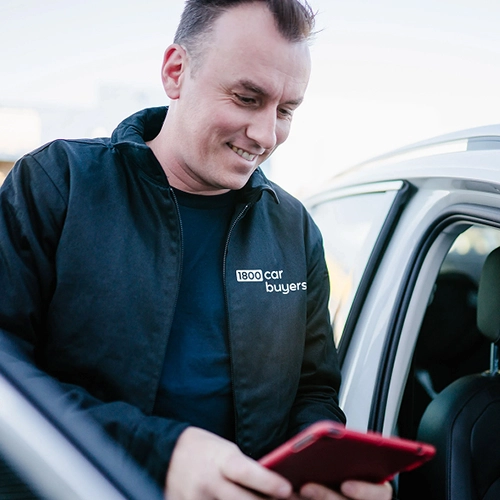 A smiling man wearing a black 1800 Car Buyers jacket looks down at a red tablet while standing next to an open car door.