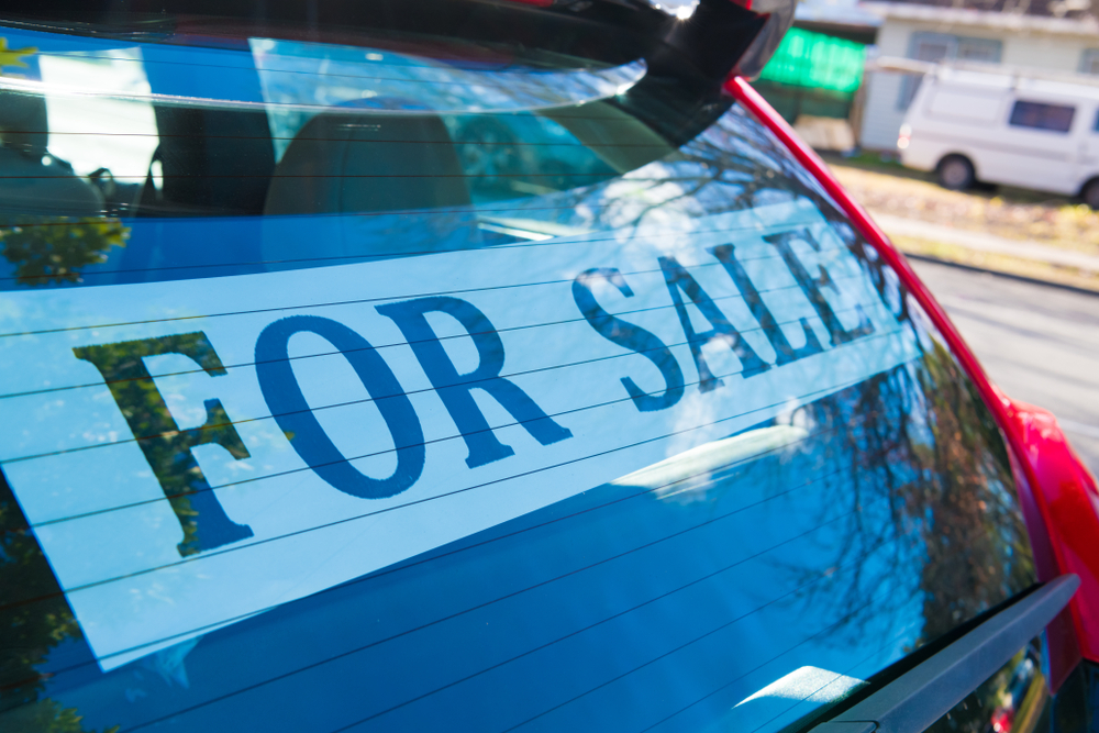 A large white "FOR SALE" sign is displayed inside the rear windscreen of a red vehicle parked on a suburban street.