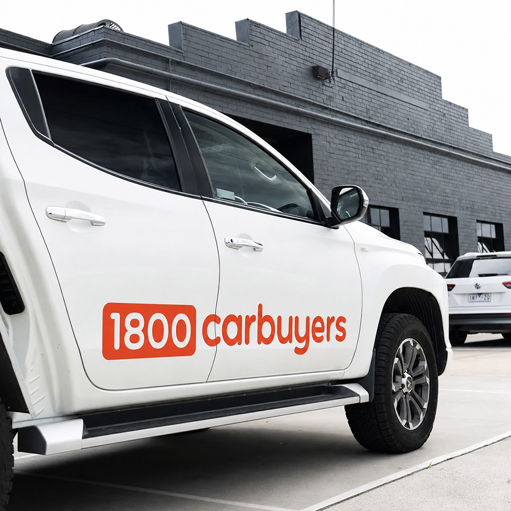 A white vehicle with orange 1800 carbuyers branding on the side is parked outside a grey multi-storey brick warehouse.