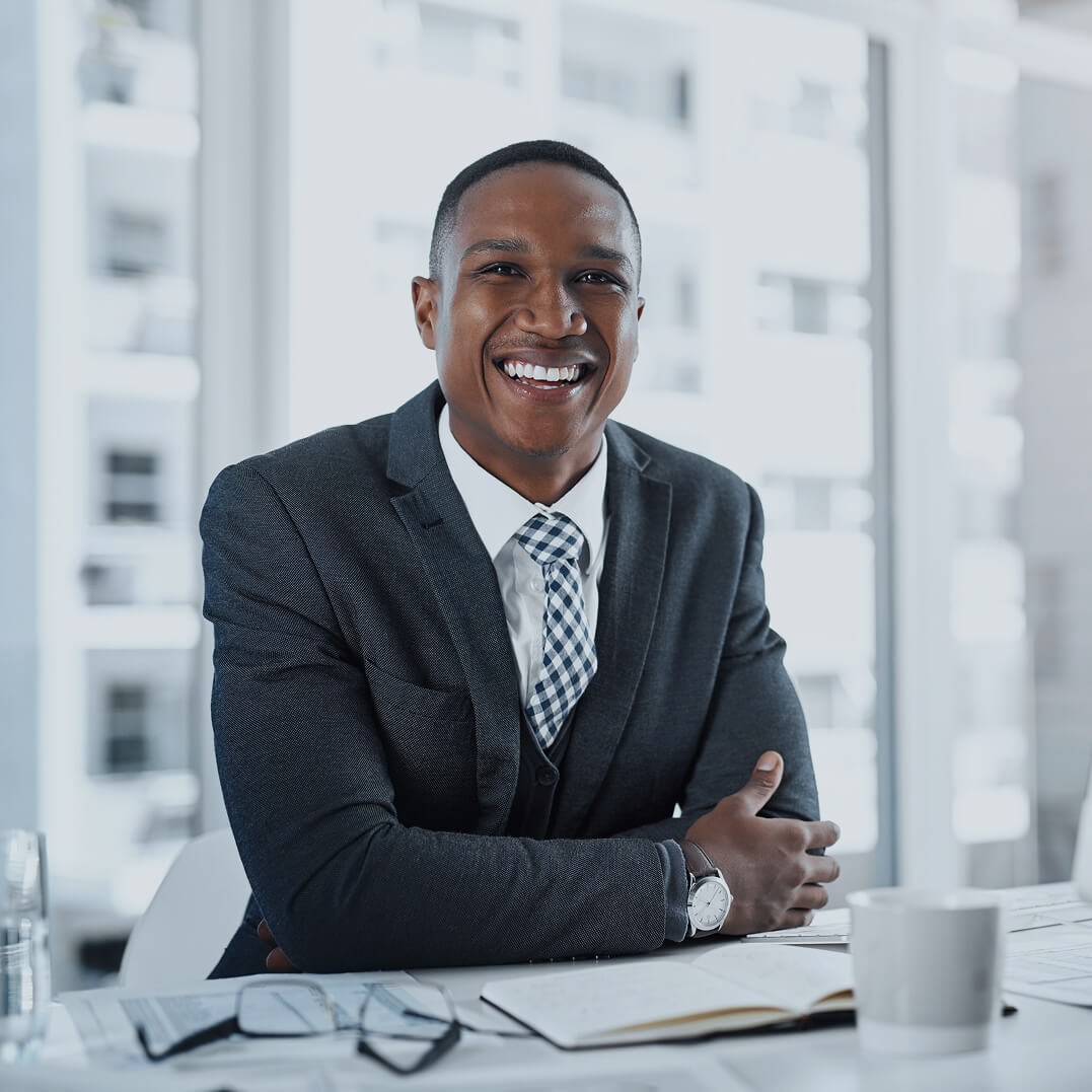 African-American male attorney sitting at his desk with high-rise buildings visible through the window behind him.