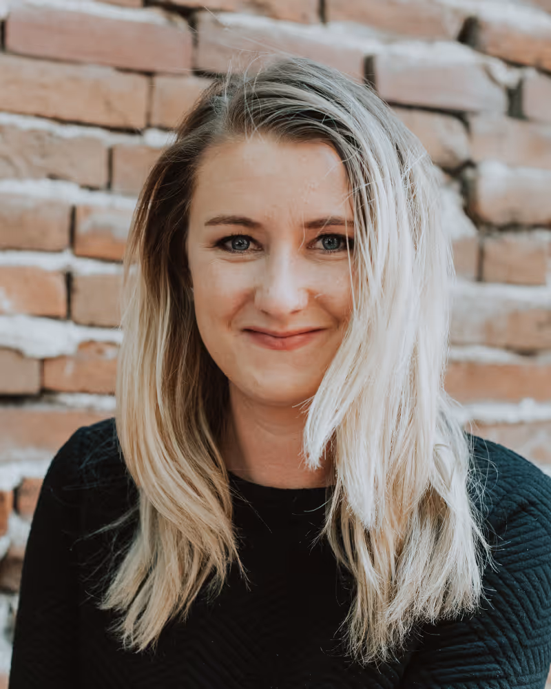 Portrait of executive director Kelsie Hower who is a smiling woman with long blonde hair wearing a black shirt in front of a brick wall.