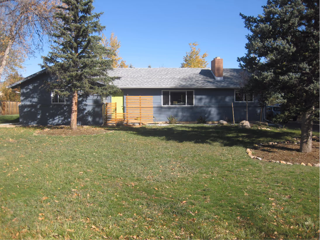 Single-story blue house with a gray roof, green front door, and wooden fence panels, surrounded by trees and green grass lawn.