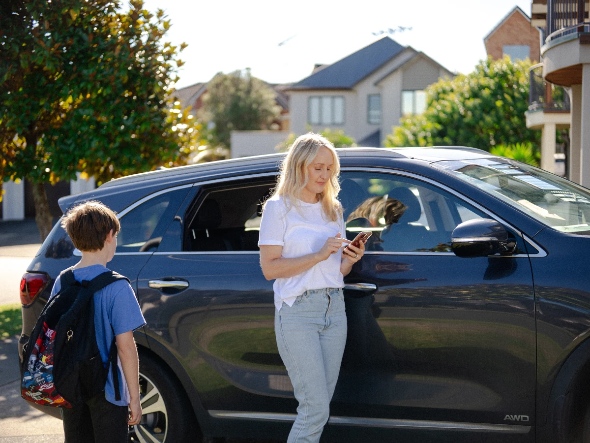 A mother in a white t-shirt uses her smartphone while standing next to a dark SUV on a sunny suburban street, as her young son stands nearby wearing a school backpack.