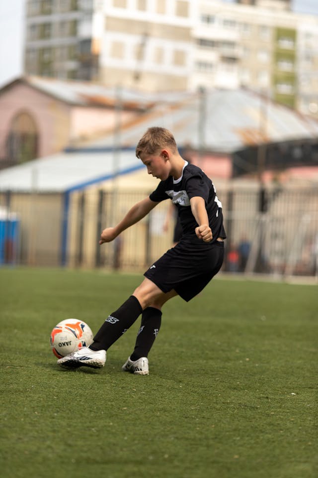 A young soccer player in black uniform striking the ball on an artificial pitch