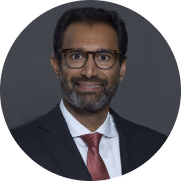 Man with glasses, beard, and dark hair wearing a suit and red tie against a gray background.