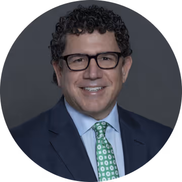 Man with curly hair and glasses wearing a blue suit, white shirt, and green patterned tie with a gray background.