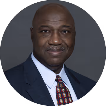 Portrait of a bald African American man in a blue suit, white shirt, and patterned tie against a gray background.