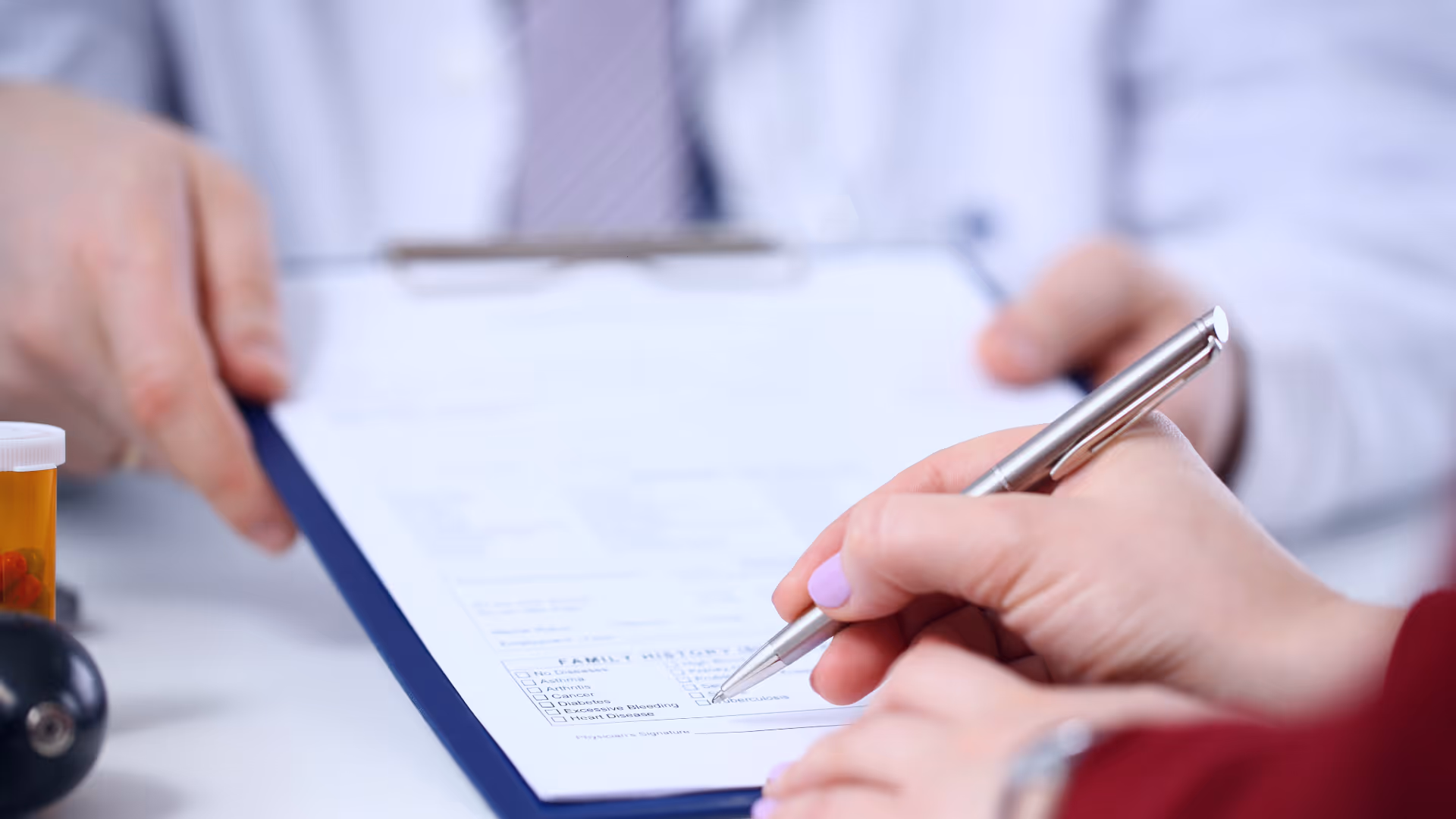A patient filling a medical form on a clipboard held by a doctor wearing a white coat and tie.