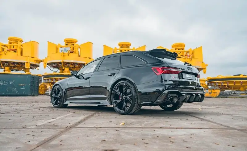 Black Audi RS6 Avant parked on a concrete surface with large yellow industrial machines in the background under a cloudy sky.