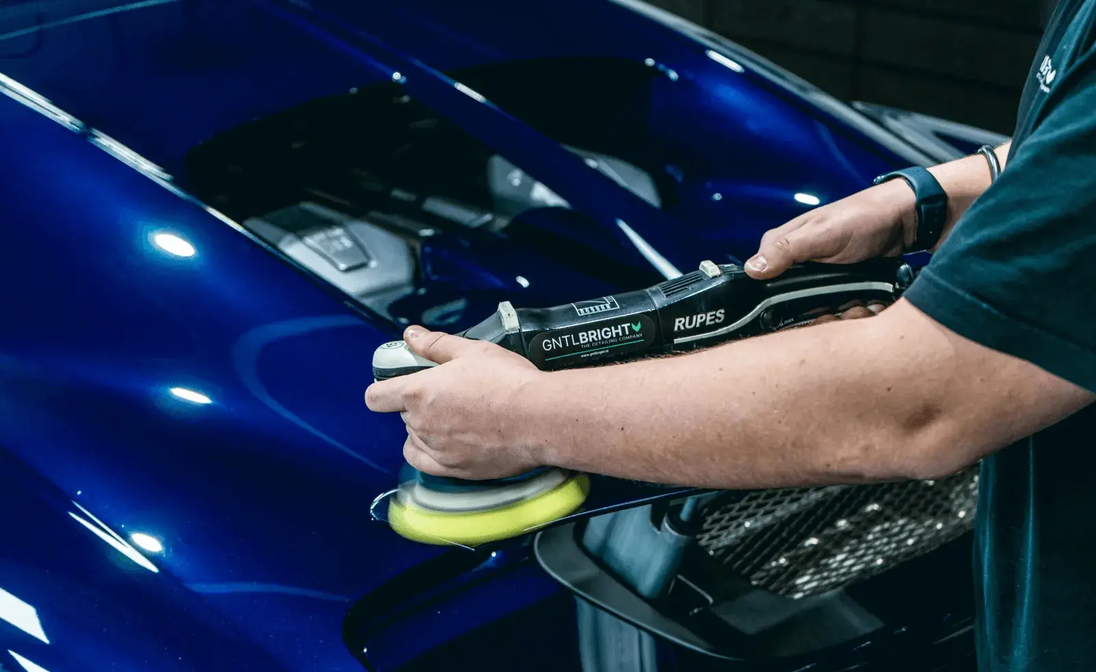 Close-up of a person using a GNTLBRIGHT Rupes polishing machine on the shiny blue hood of a car with the engine visible underneath the open hood.