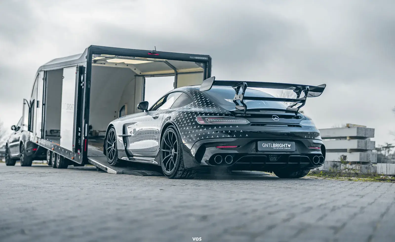 Black Mercedes AMG GT sports car with a large rear spoiler and patterned exterior being loaded onto an enclosed car trailer attached to a pickup truck on a paved surface.