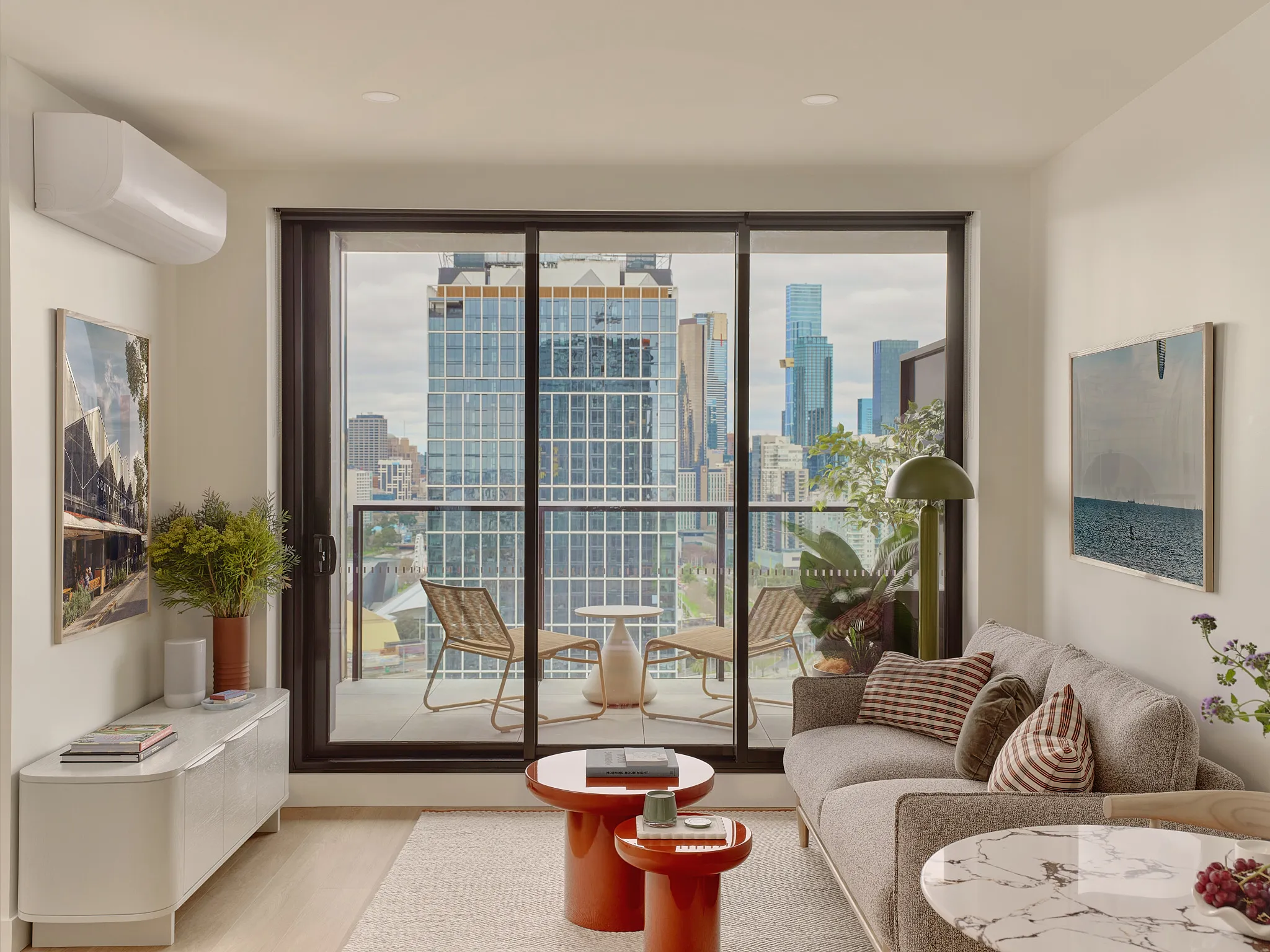 Modern living room with large sliding glass doors opening to a balcony with two wicker chairs and a small white table overlooking a cityscape with tall buildings. Inside, there is a gray sofa with striped pillows, a pair of red round coffee tables, a white TV console with books and a vase of greenery, and a round marble-top table with fruit. The walls have framed photographs, and a green floor lamp stands near the sofa.