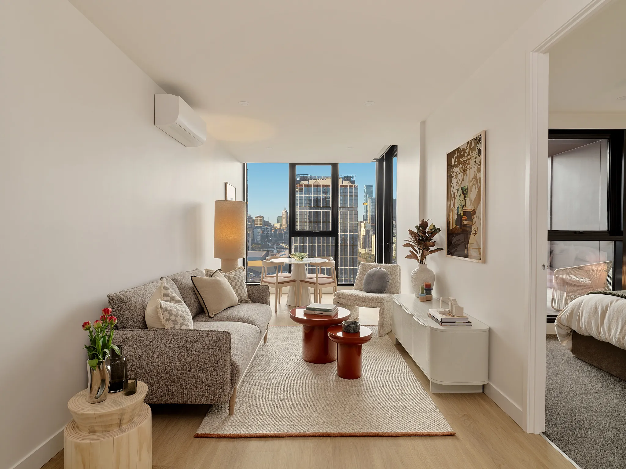 Modern living room with light wood floor, gray sofa with pillows, side table with flowers, two round reddish coffee tables, a white chair, a floor lamp, a white media console with decor, and large windows showing city skyscrapers in daylight; bedroom visible through open door on the right.