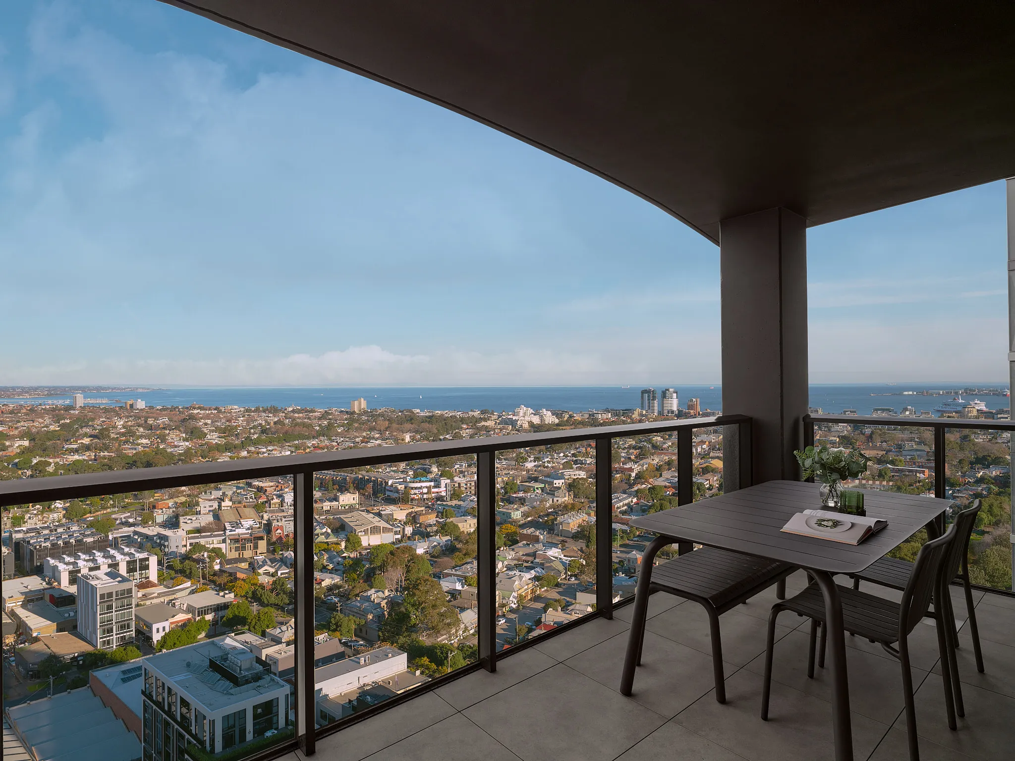 Modern balcony with glass railing overlooking a cityscape and ocean under a blue sky, furnished with a table, two chairs, and a bench with a book and a flower vase on the table.