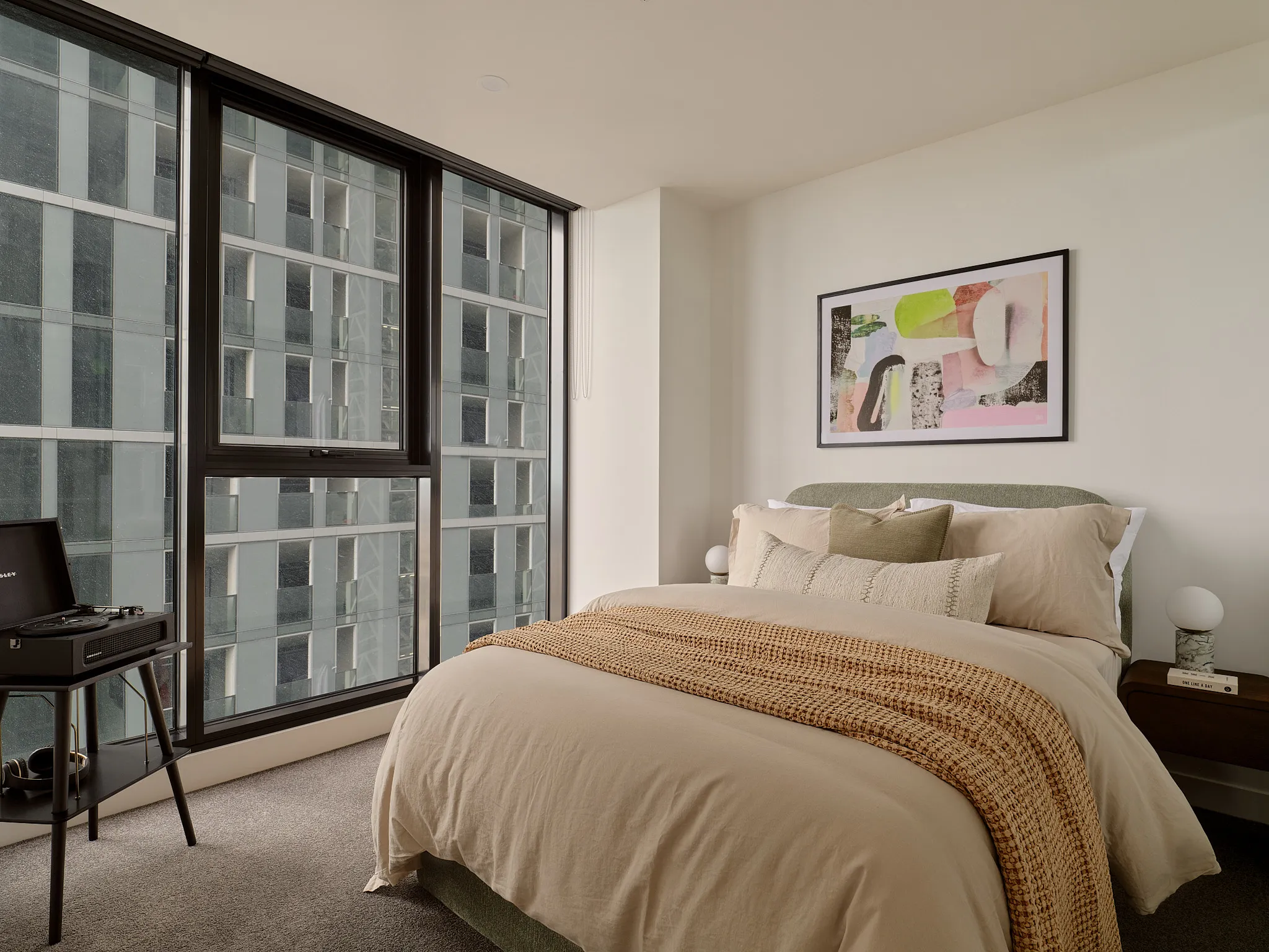 Modern bedroom with large window, beige bedding, textured throw, and abstract artwork above bed.