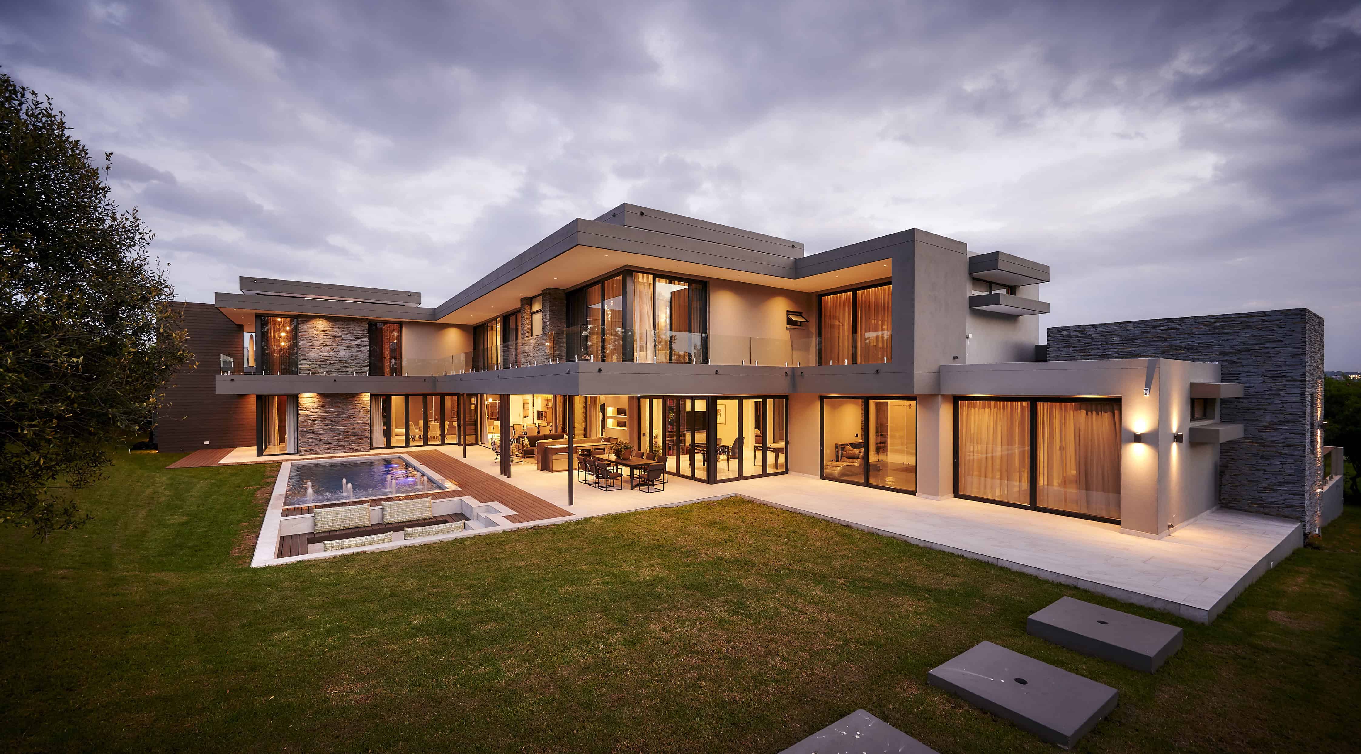 Modern multi‑story home with expansive lawn, large windows and warm evening lighting designed by Interior Designer Johannesburg.