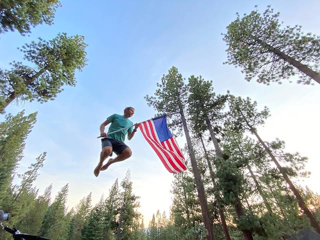 Photo of Tommy Caldwell barefoot and leaping in a conifer grove, holding an American flag.
