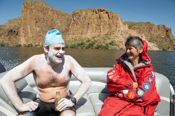 Joe Zemaitis and Claire Russell sitting on a boat. Russell is wearing a red parka decorated with successful swims.