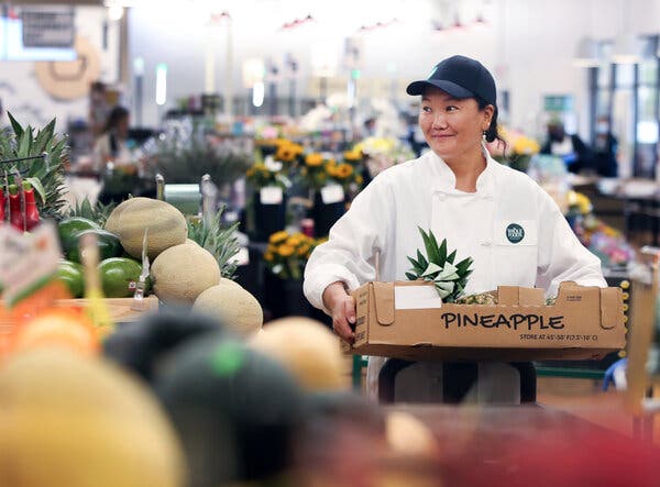 Lhakpa Sherpa in a white Whole Foods uniform and blue cap carries a box of produce.