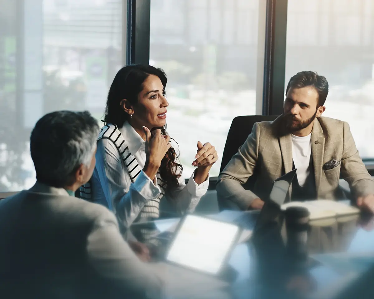 Three business partners engaged in a discussion around a conference table in a modern office.