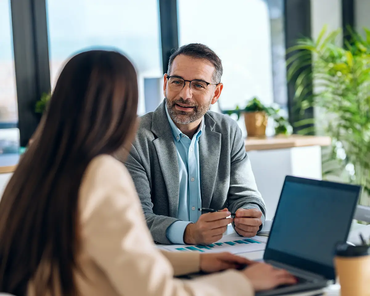 Accountant wearing glasses and gray blazer talking to a woman with long brown hair in an office setting with a laptop and documents on the table.