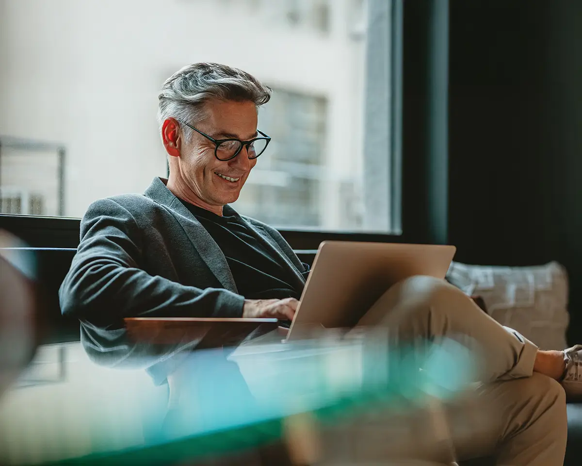Smiling man with gray hair and glasses sitting comfortably using a laptop near a window.