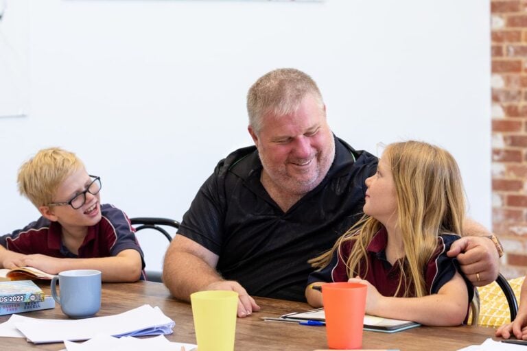 A dad with two kids sitting down at the dining table, looking at each other smiling. The kids are wearing school uniform and doing homework at the table.