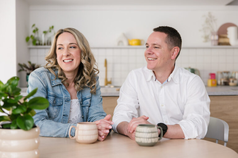 MyBudget clients Erin and Adam sitting at a table with mugs and smiling because they have no more money worries.