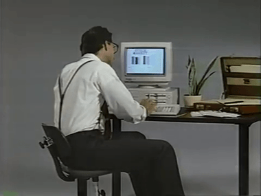 Man with suspenders seated at a desk working on an old desktop computer with a small plant and an open briefcase nearby.