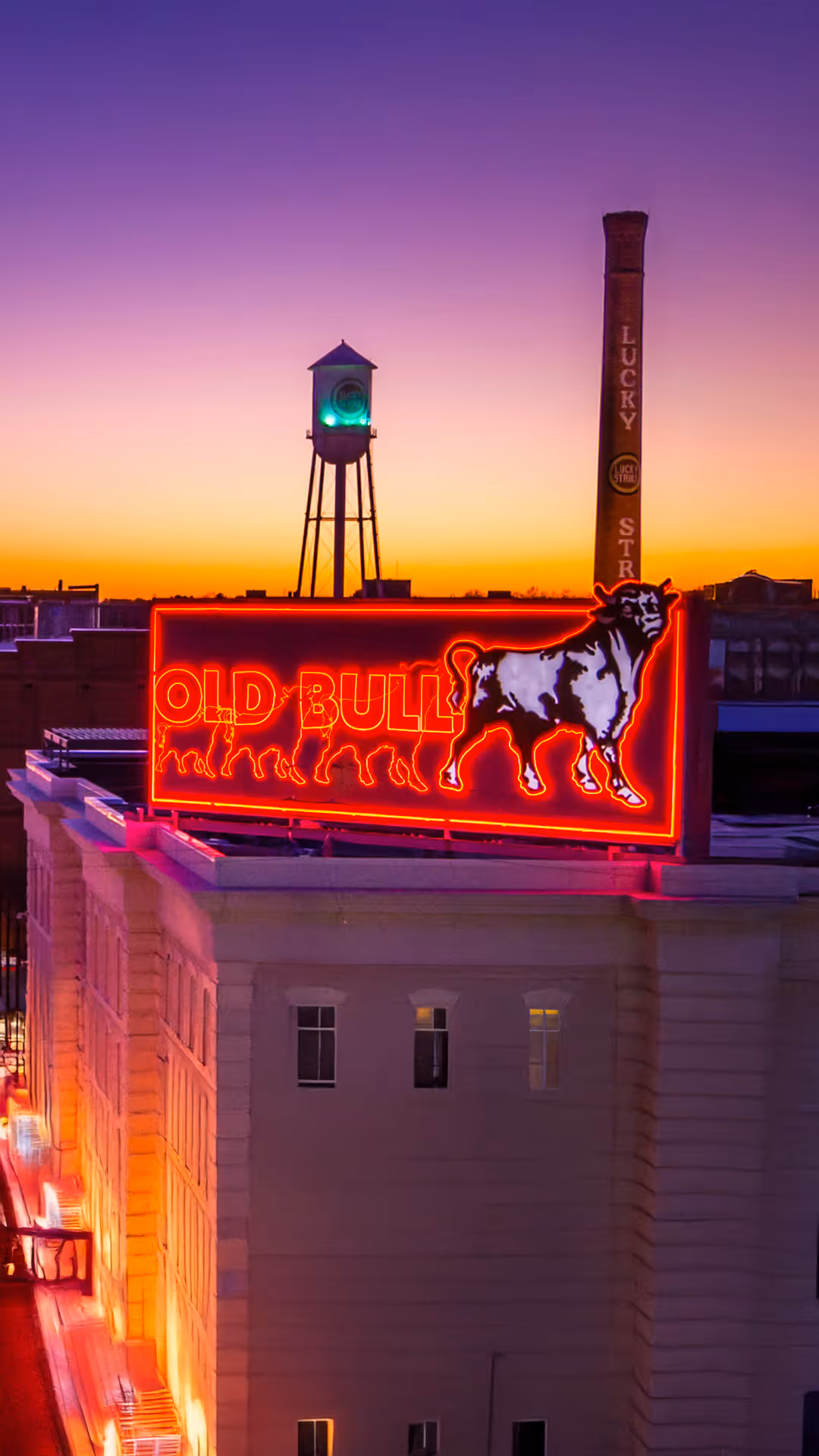 Rooftop neon sign of a bull with the text 'OLD BULL' glowing red against a sunset sky, with a water tower and a tall chimney marked 'LUCKY STRIKE' in the background.