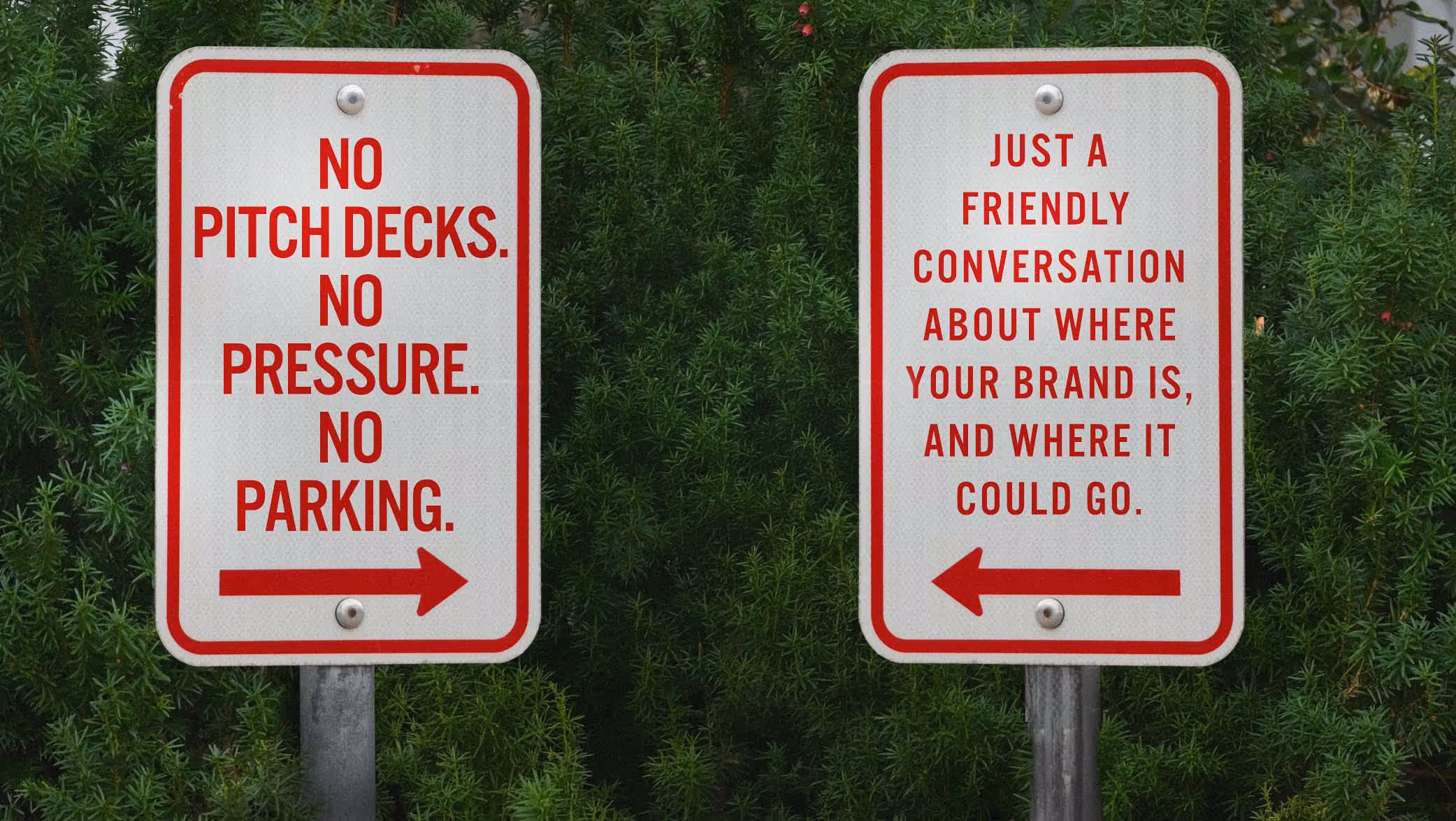 Two white signs with red borders and text on poles in front of green bushes; left sign reads 'No pitch decks. No pressure. No parking.' with arrow pointing right; right sign reads 'Just a friendly conversation about where your brand is, and where it could go.' with arrow pointing left.