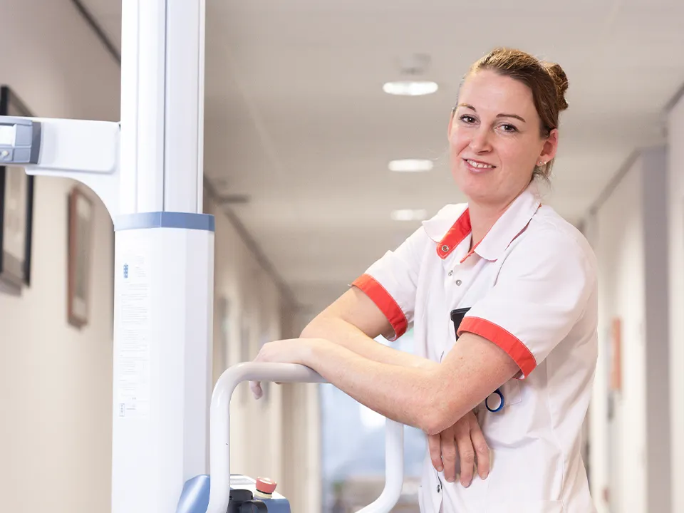 Lachende vrouwelijke zorgverlener, leunend tegen medische apparatuur in een ziekenhuisgang.