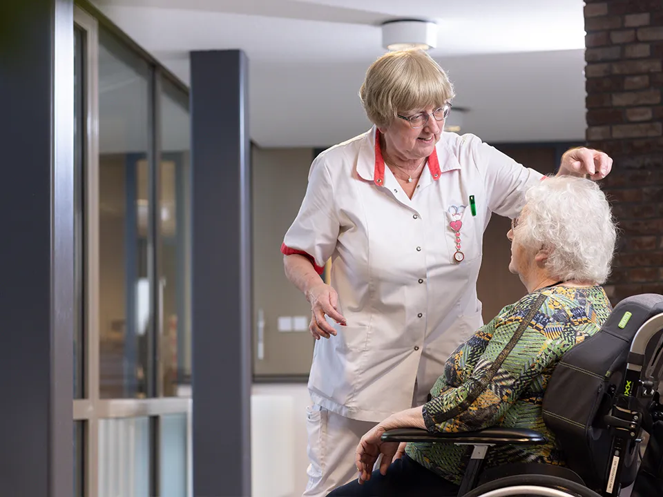 Zorgmedewerker in wit uniform die zachtjes het hoofd aanraakt van een oudere vrouw in een rolstoel in een zorginstelling.
