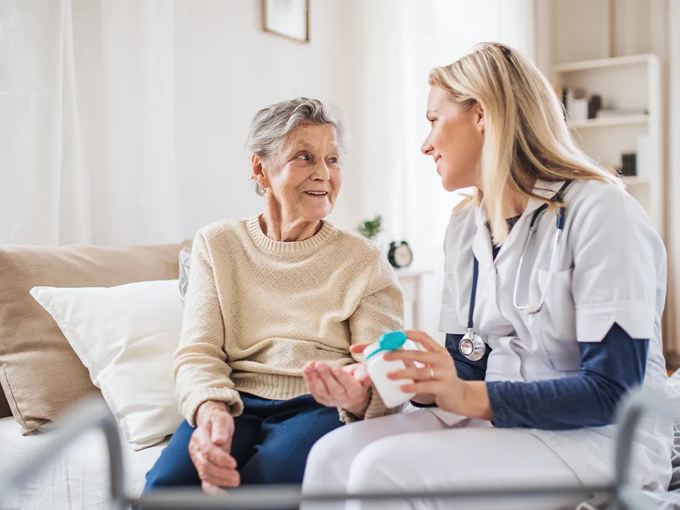 Bejaarde dame op de bank, glimlachend naar een verpleegster met een flesje medicijnen.
