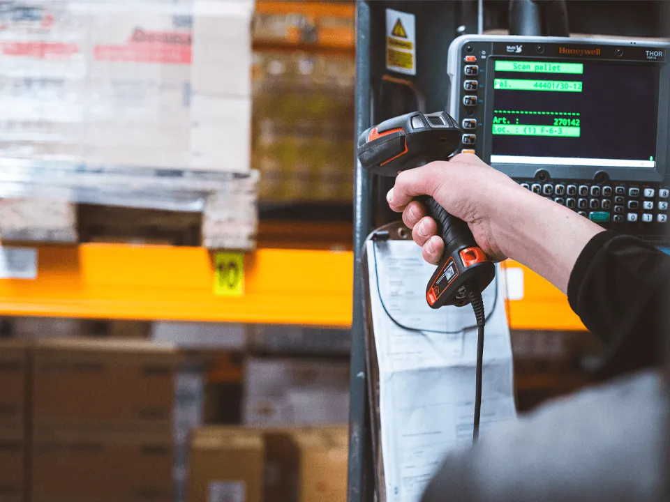 Hand die een barcodescanner vasthoudt in een magazijn, scant een pallet met schappen en dozen op de achtergrond.