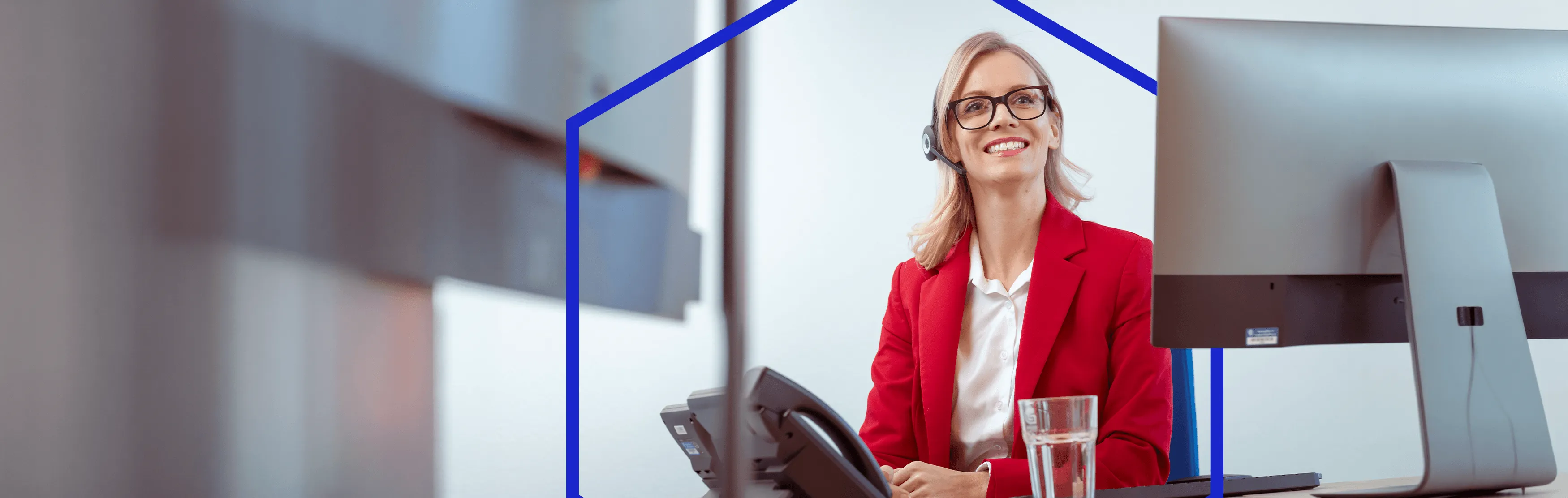 Vrouw met bril en headset glimlacht terwijl ze achter een bureau zit met een telefoon, glas water en een computerscherm.