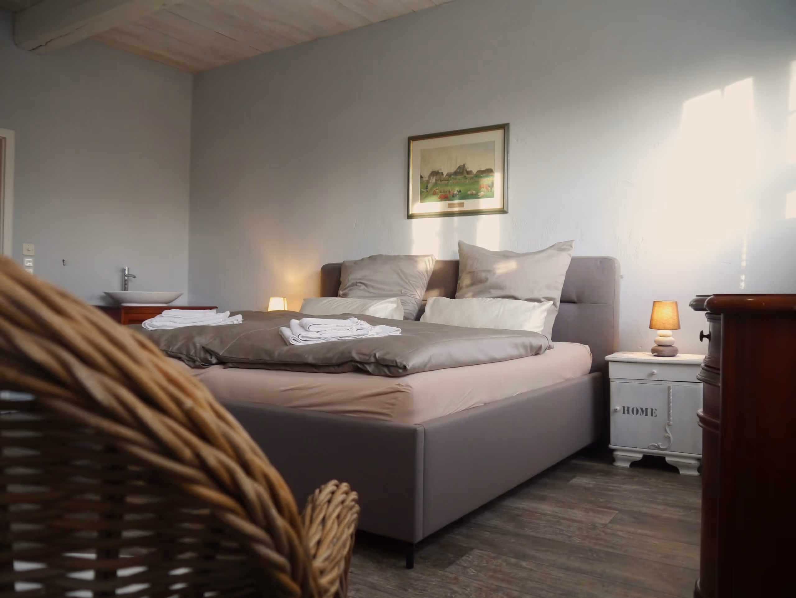 Hell eingerichtetes Schlafzimmer mit einem grauen Bett, Kissen, weißen Handtüchern auf der Bettdecke, Nachttisch mit Lampe und Korbsessel im Vordergrund.